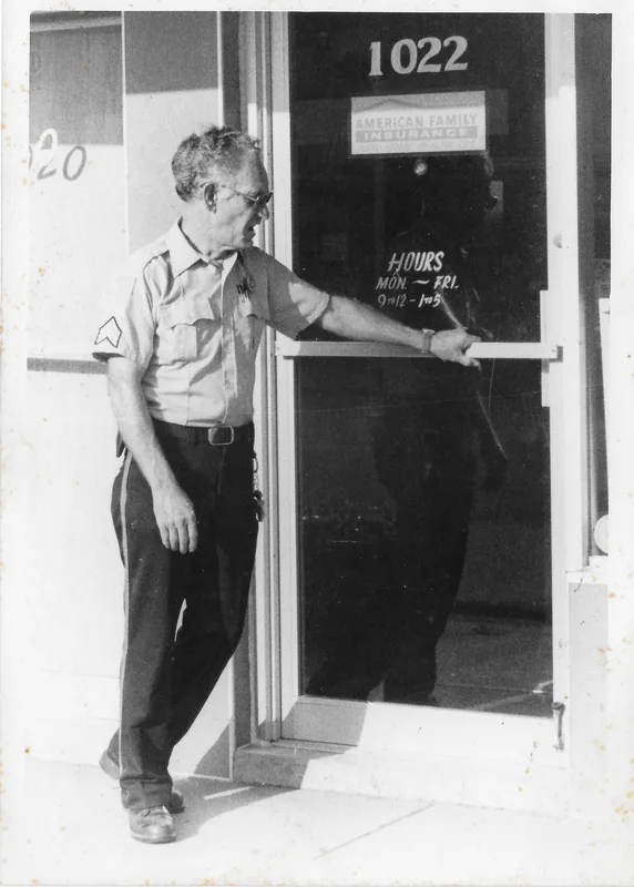 A man in a uniform shirt with sergeant stripes leans on a glass door with an American Family Insurance sign