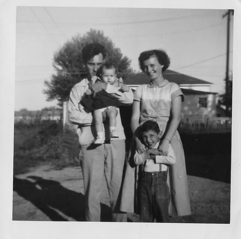 A man holding a baby and a woman with her hand on a young boy's shoulder, standing in front of a house, in black and white