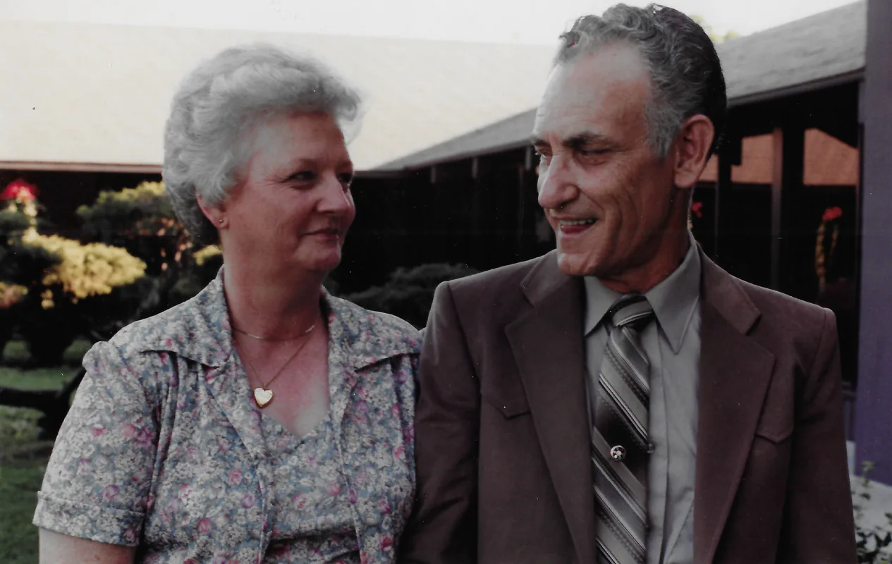 A smiling older woman in a floral blouse and a man in a brown jacket and striped tie look at each other outdoors