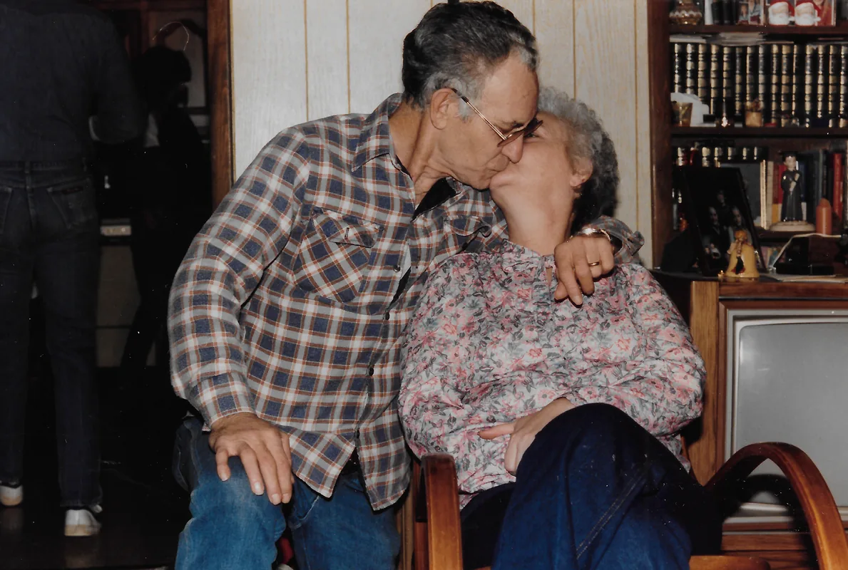 An older man in a plaid shirt kisses an older woman sitting in a rocking chair