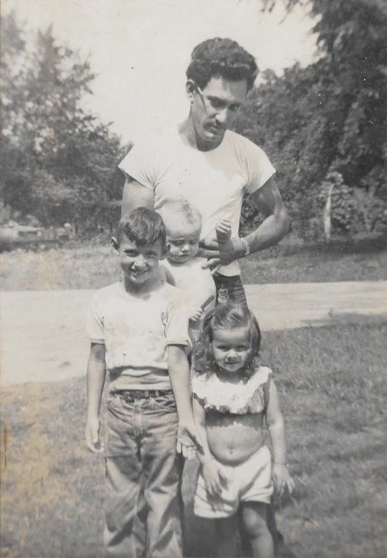 A man in a white T-shirt stands behind three young children in a yard, in an old black-and-white photograph