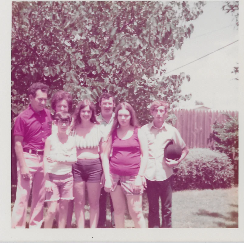 Seven people stand in a row in a backyard in front of a tree and wooden fence, in a 1970s color photograph